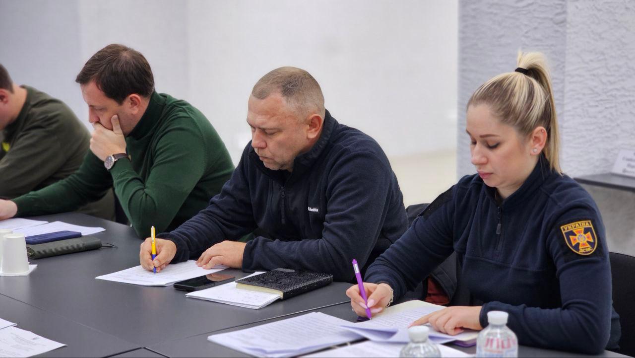 Крупний план трьох учасників наради, які зосереджено роблять записи за столом. Жінка праворуч одягнена у темну флісову кофту з нашивкою: УКРАЇНА та емблемою, схожою на символіку ДСНС. Поруч із нею сидять двоє чоловіків у цивільному одязі.