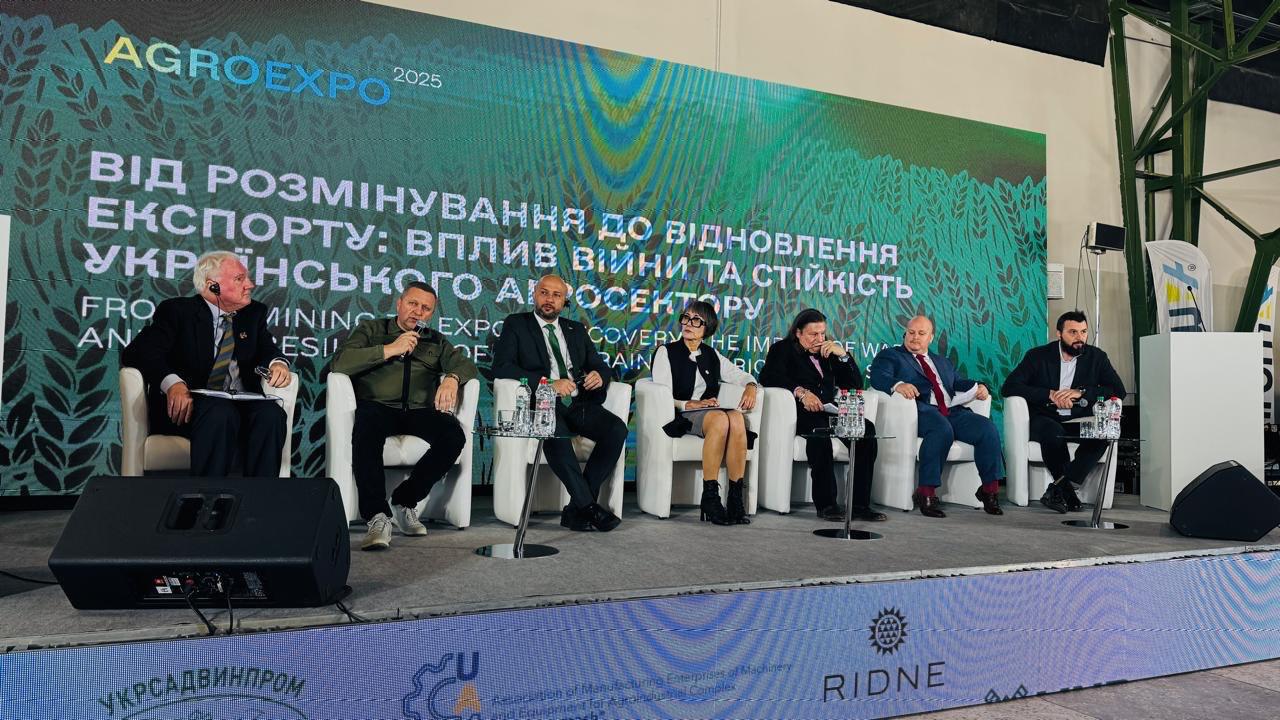Панельна дискусія на сцені конференції AGROEXPO 2025, де семеро учасників (шість чоловіків і одна жінка) сидять на кріслах. На великому екрані позаду них відображено тему дискусії українською та англійською мовами: ВІД РОЗМІНУВАННЯ ДО ВІДНОВЛЕННЯ ЕКСПОРТУ: ВПЛИВ ВІЙНИ ТА СТІЙКІСТЬ УКРАЇНСЬКОГО АГРОСЕКТОРУ.