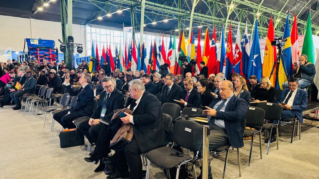 Велика група учасників, одягнених у діловий одяг, сидить на стільцях під час міжнародної конференції чи форуму. На задньому плані видно довгий ряд прапорів багатьох країн, що підкреслює міжнародний характер події.