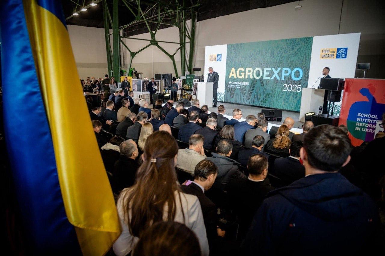 Широкий план великої аудиторії, яка сидить на конференції AGROEXPO 2025. На сцені чоловік виступає за трибуною перед великим екраном з логотипом AGROEXPO. На передньому плані ліворуч — частина великого українського прапора.