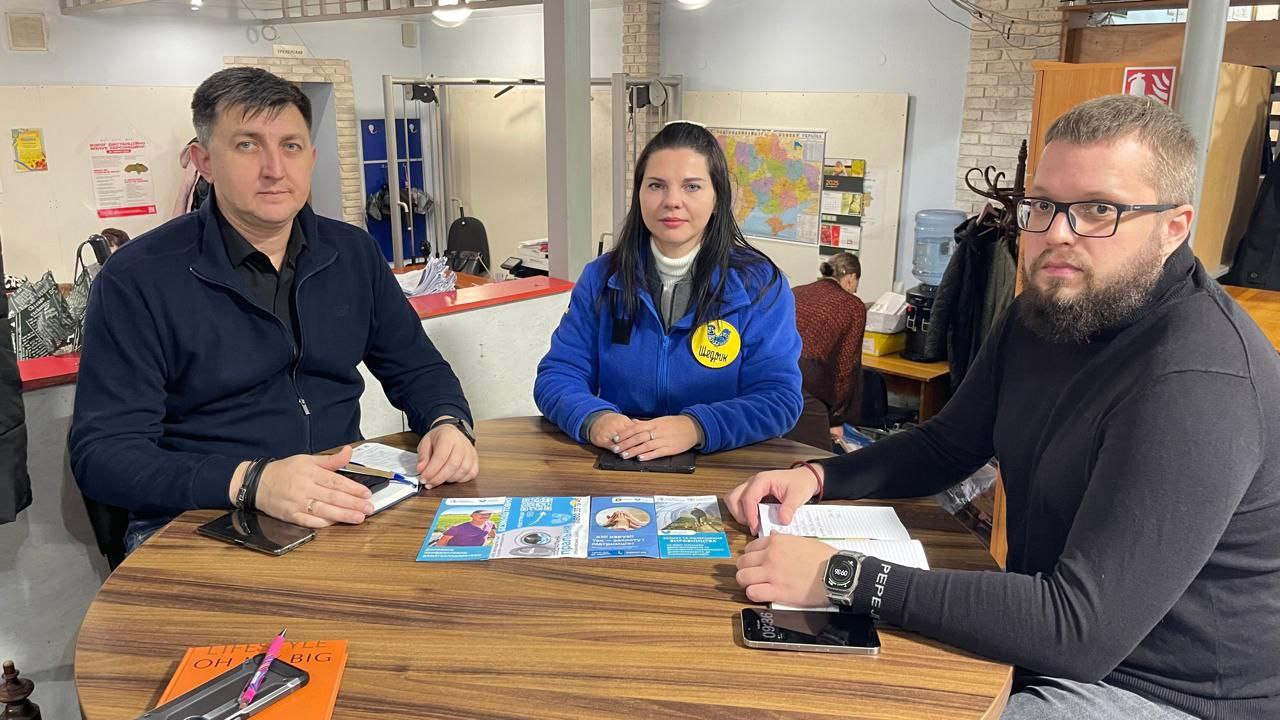 Три особи – двоє чоловіків і жінка в синій кофті з логотипом – сидять за круглим дерев'яним столом під час зустрічі. На столі лежать документи та інформаційні матеріали.