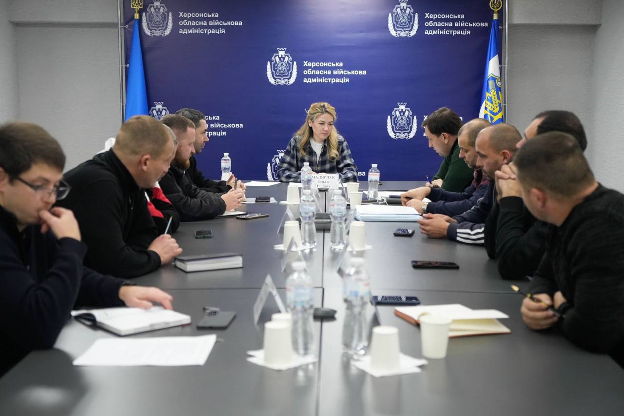 Нарада за довгим столом, на якій присутні близько десяти чоловіків та одна жінка в центрі. На задньому плані — синій банер з логотипом та написом: Херсонська обласна військова адміністрація та державний прапор України.