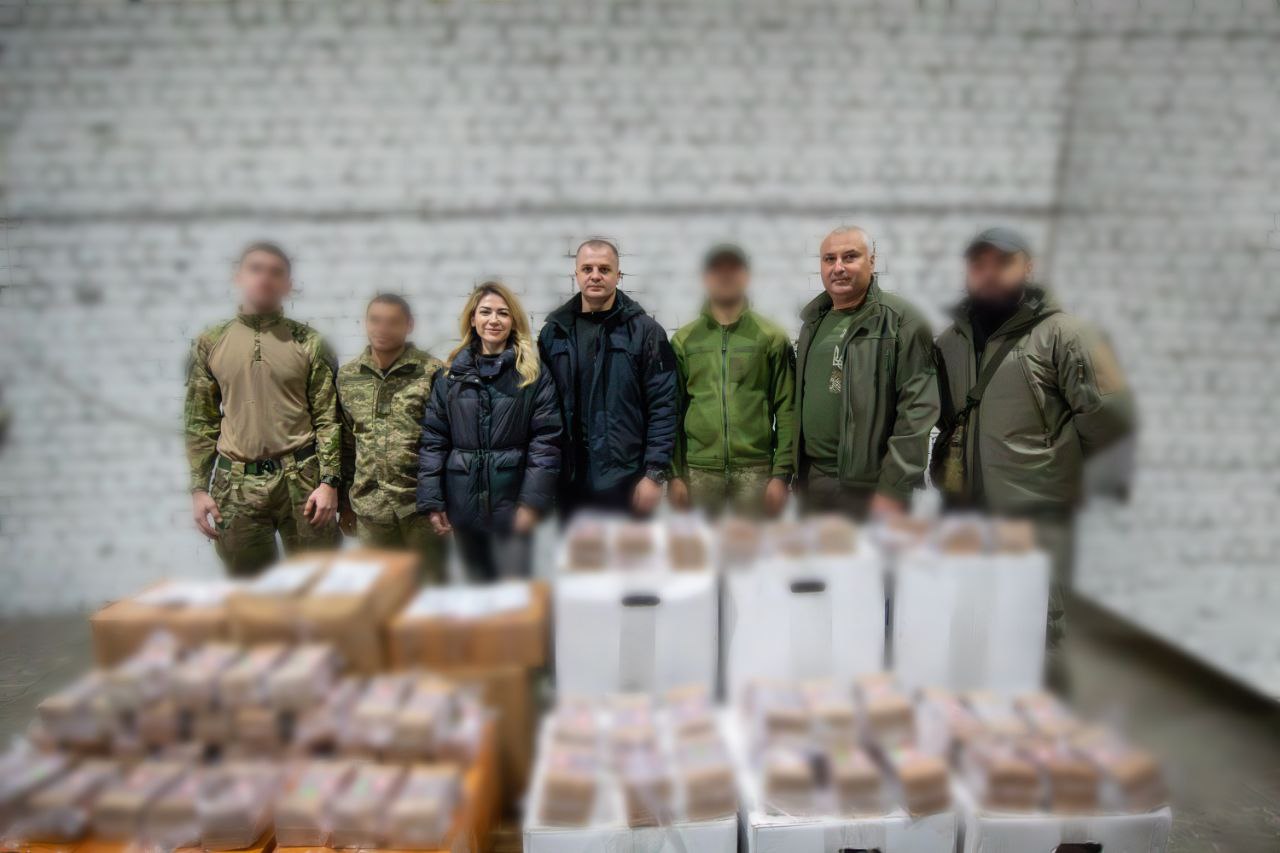 Група з шести осіб (чотири чоловіки у військовій або тактичній формі та жінка з чоловіком у цивільному одязі) стоять на складі перед великою кількістю коробок та палет з гуманітарною допомогою або вантажем. Обличчя деяких людей розмиті.