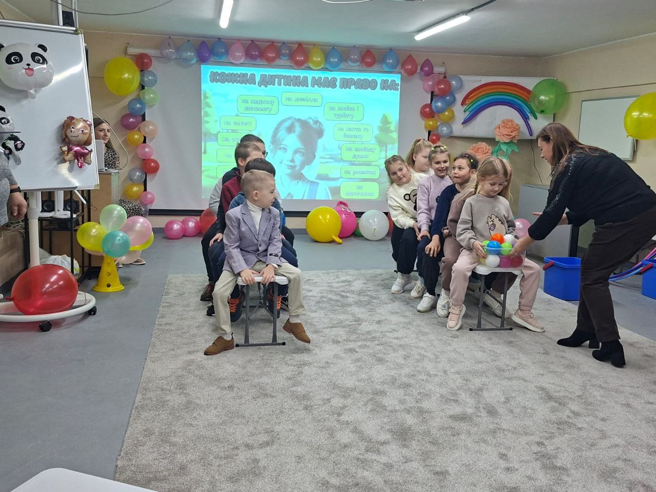 Група дітей сидить у ряд на стільцях у прикрашеному повітряними кульками приміщенні, беручи участь у занятті чи грі під керівництвом дорослої жінки. На екрані позаду них відображено тему заходу: КОЖНА ДИТИНА МАЄ ПРАВО НА....