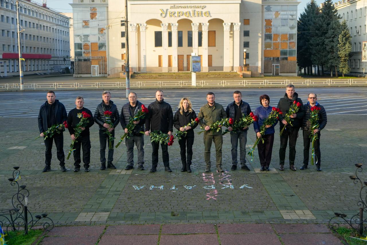 Група людей (чоловіки та жінка) стоїть у ряд із букетами червоних троянд перед пошкодженою будівлею кінотеатру «Україна» на площі.
