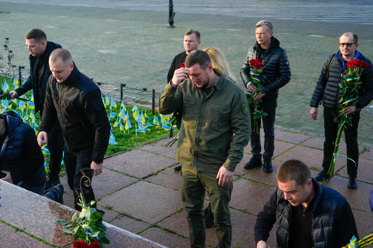 Група чоловіків стоїть біля меморіальної плити, покладаючи червоні троянди; деякі з них мають скорботний вираз обличчя. Позаду видно багато маленьких українських прапорів.