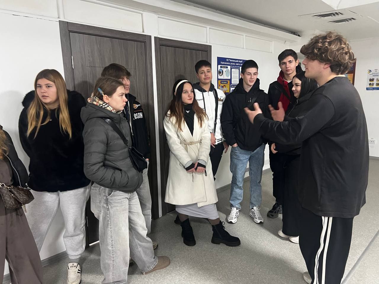 Група з восьми-десяти підлітків та молодих людей стоїть у коридорі біля дверей, уважно слухаючи високого хлопця з кучерявим волоссям, який активно жестикулює під час розмови.