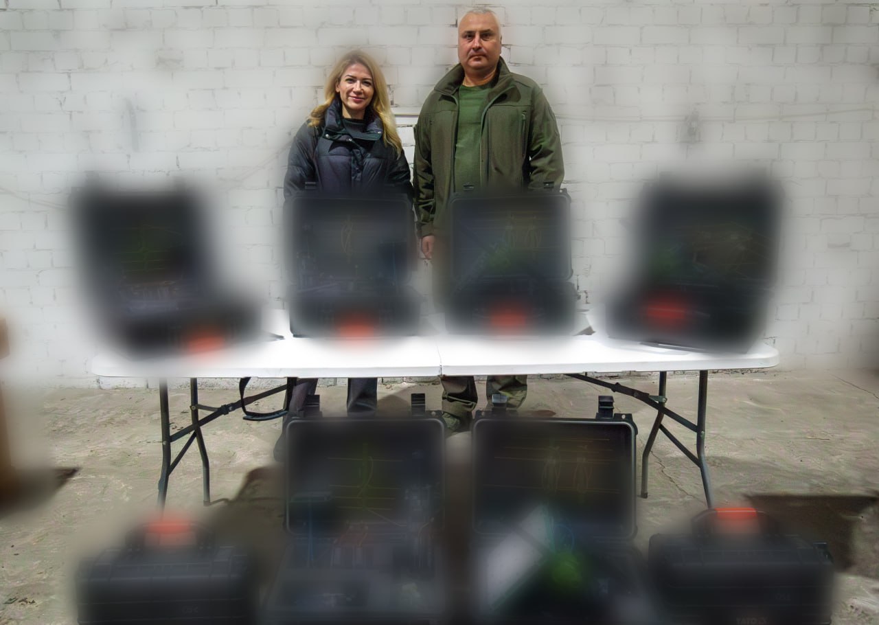 Жінка та чоловік стоять позаду столу з кількома чорними кейсами, що стоять відкритими, всередині яких видно якесь електронне обладнання (з розмитими деталями). Вони посміхаються. За ними — біла цегляна стіна.