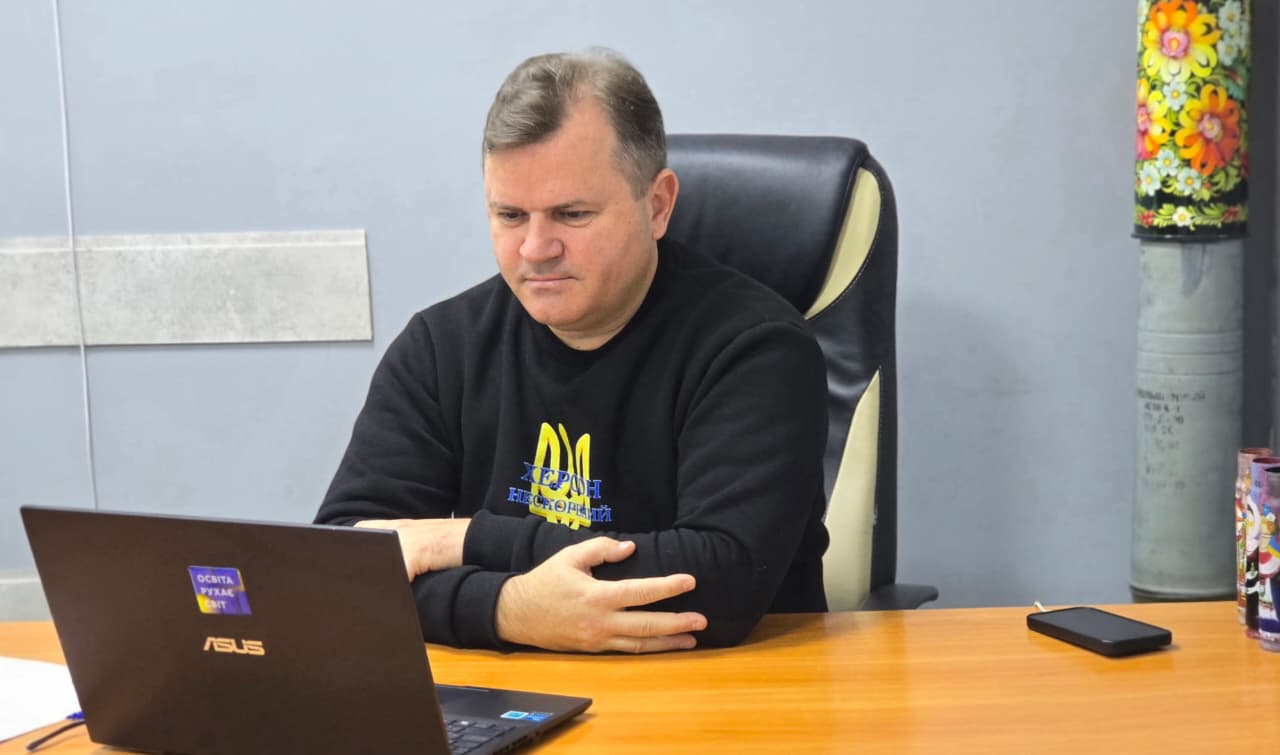 Чоловік середнього віку в чорному світшоті з жовто-синім тризубом на грудях сидить за офісним столом, дивлячись на екран ноутбука ASUS перед собою. На столі також лежить мобільний телефон і стоїть розписаний квітковим візерунком відпрацьований снаряд або гільза.