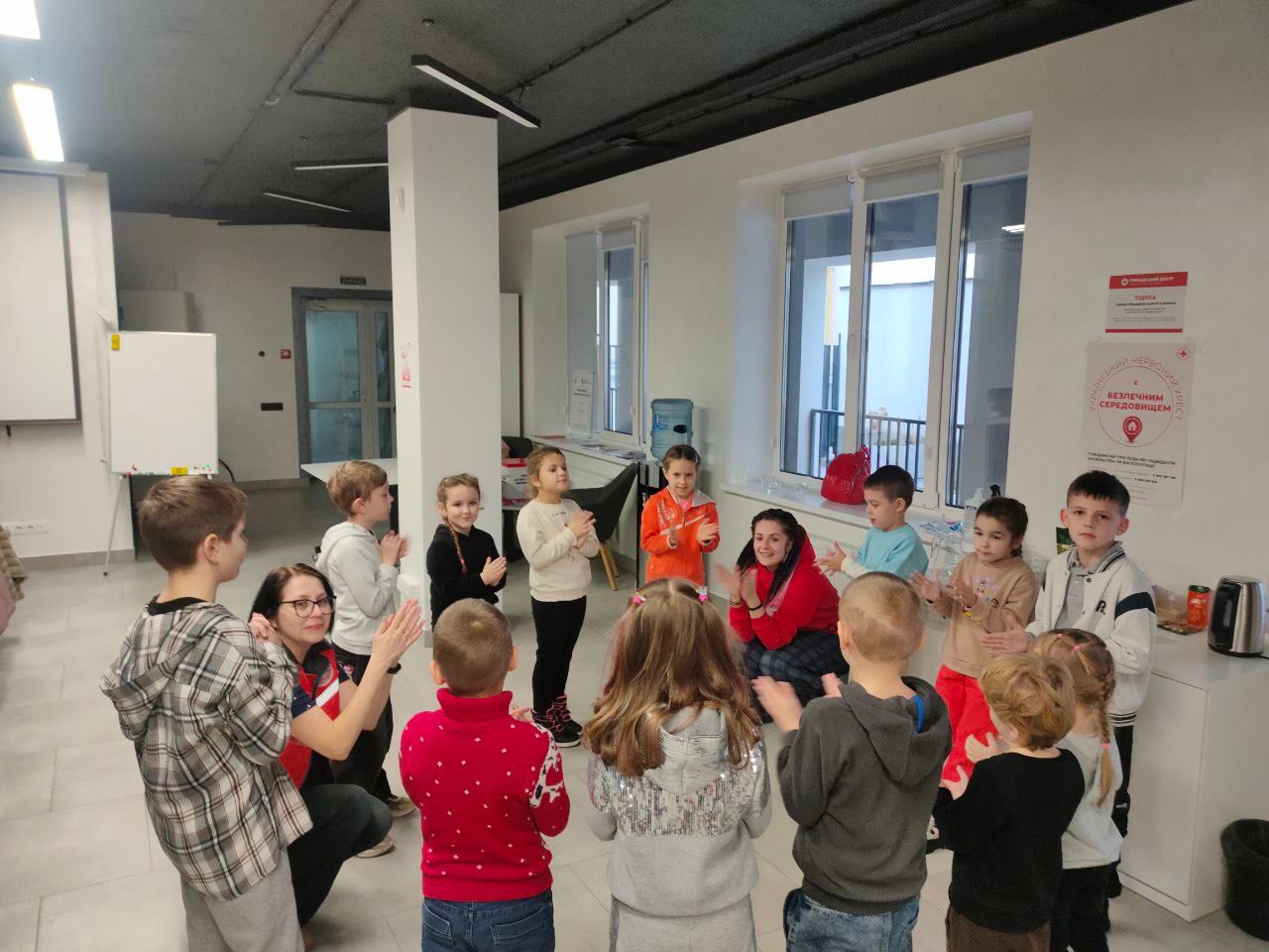 Група дітей стоїть півколом навколо двох дорослих у світлій, сучасній кімнаті з великими вікнами. Всі учасники плескають у долоні, імовірно, граючи в якусь гру чи беручи участь у занятті. На стіні позаду видно логотип Червоного Хреста та напис: Безпечним середовищем.