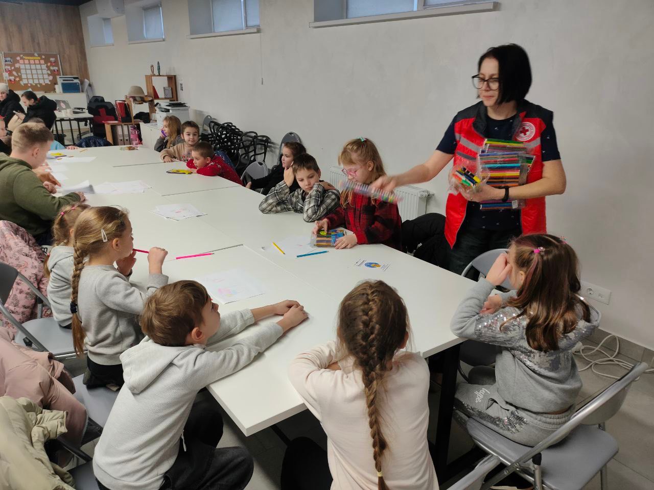Група дітей сидить за довгим білим столом у приміщенні, займаючись малюванням. Жінка-інструктор у червоному жилеті Червоного Хреста стоїть праворуч і пропонує дітям набір кольорових олівців/фломастерів. Діти зосереджені на своїх аркушах паперу.