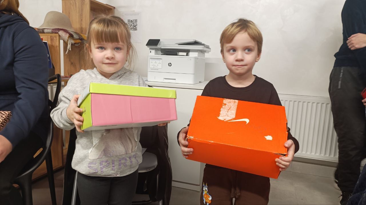 Двоє маленьких дітей, дівчинка та хлопчик, стоять поруч і тримають у руках коробки. Дівчинка тримає рожево-салатову коробку, а хлопчик — помаранчеву коробку з логотипом Nike. Діти дивляться прямо в об'єктив. На задньому плані видно офісну техніку та дерев'яні полиці.