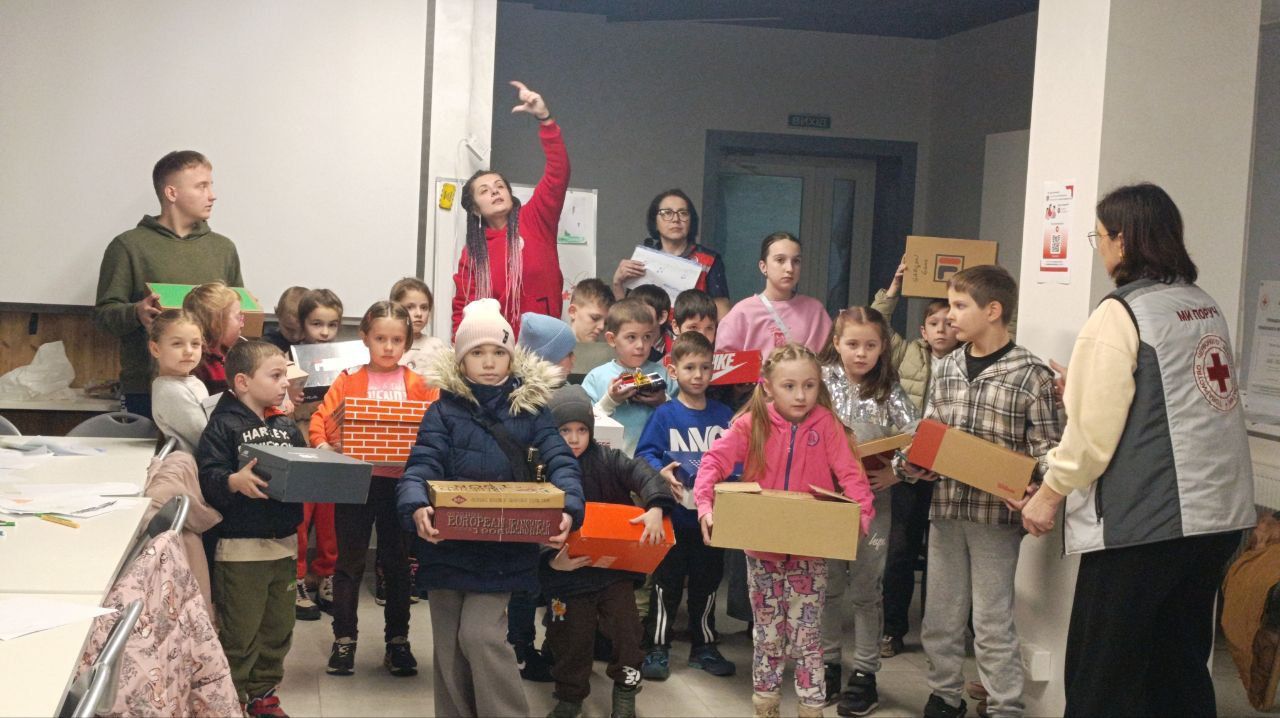 Велика група дітей стоїть разом у приміщенні, більшість із них тримають у руках подарункові коробки різних кольорів та розмірів (ймовірно, від взуття). Поруч із дітьми видно двох жінок-організаторів: одна в червоній куртці, яка щось показує рукою, інша – у сірому жилеті з логотипом Червоного Хреста.