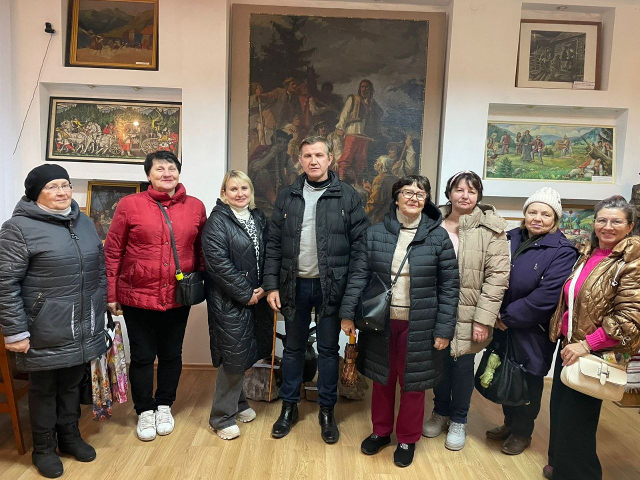 Група з восьми дорослих людей (одного чоловіка та семи жінок) у зимовому одязі стоїть у приміщенні художньої галереї, позуючи перед великою картиною та оточені меншими картинами на стінах.