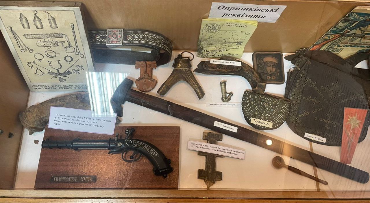 Виставка музейних експонатів у скляній вітрині, підписана як: Опришківські реквізити, що містить різноманітні предмети, пов'язані з опришками, зокрема старовинний пістолет XVIII ст., тобівка (сумка для куль), шкіряний пояс із металевою пряжкою, ремені, металеві хрести, елементи зброї та інше приладдя.