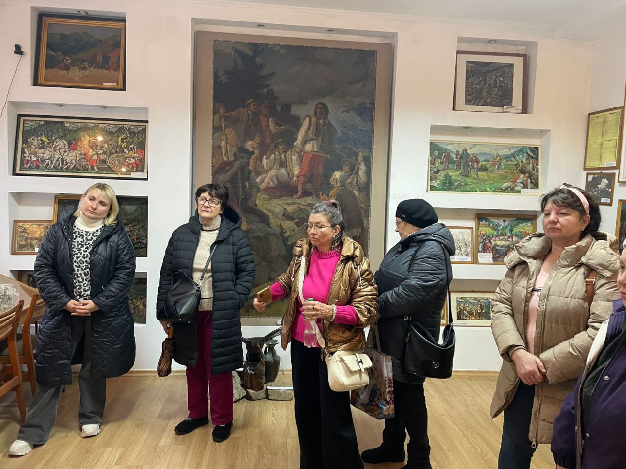 Група із п'яти жінок стоїть у художній галереї, розглядаючи велику вертикальну картину на задньому плані, оточену багатьма меншими картинами на стінах.