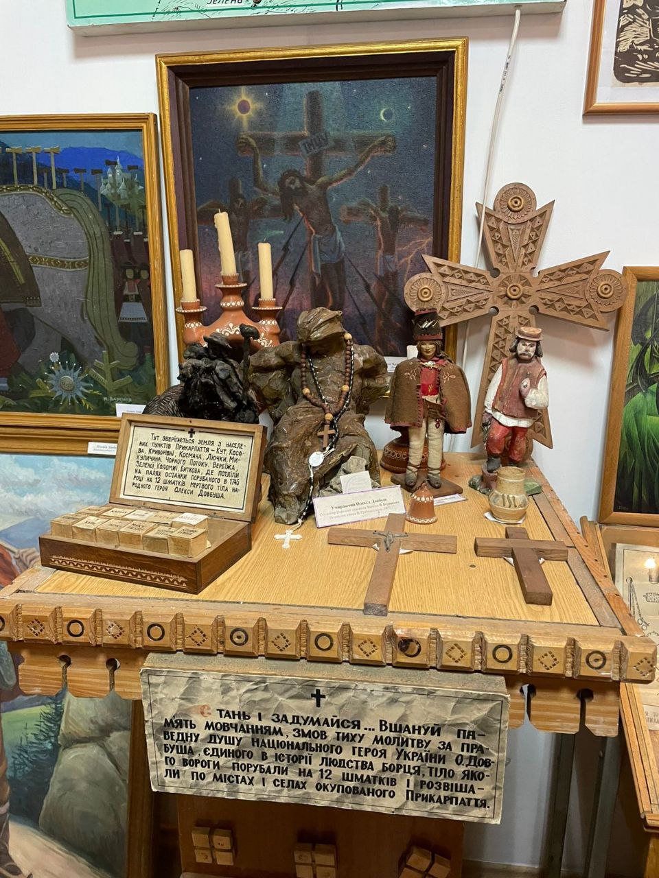 Експозиція в музеї на дерев'яному столі, що містить релігійні та етнографічні артефакти, включаючи дерев'яні різьблені хрести, свічники, невеликі фігурки, що зображують гуцулів, скульптуру чоловіка в молитві з вервицею, а також таблички з пояснювальним текстом, на задньому плані видно картину із зображенням Розп'яття.