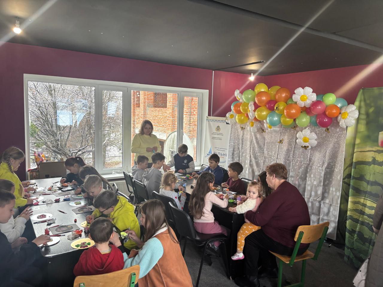  Група дітей та кілька дорослих займаються творчістю (малюванням чи виготовленням поробок) за столами у світлій кімнаті з великим вікном та яскравим оформленням, прикрашеною гірляндою з повітряних кульок.