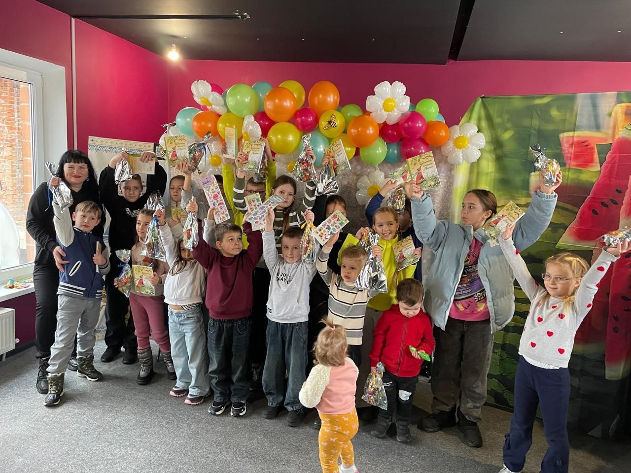 Групова фотографія дітей різного віку та двох дорослих у яскраво оформленій кімнаті (рожеві стіни, фон з повітряних кульок та зображенням кавуна), які радісно тримають над головою подарунки.