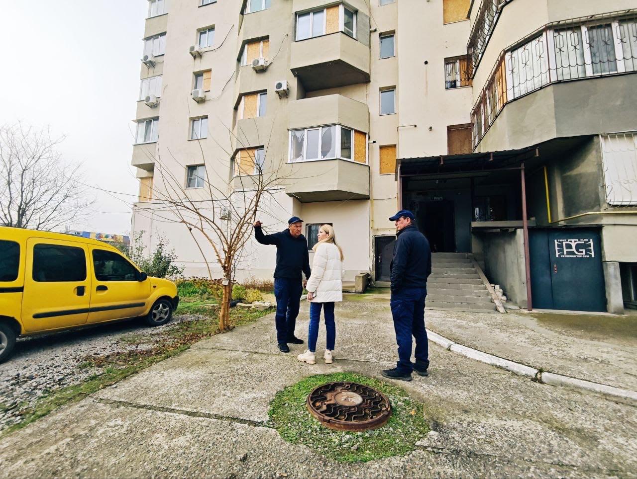 Три людини (двоє чоловіків у темному одязі та жінка у білій куртці) стоять і спілкуються біля входу в багатоквартирний будинок, вікна якого закриті захисними щитами (фанерою) через пошкодження. На передньому плані видно жовтий фургон та каналізаційний люк.