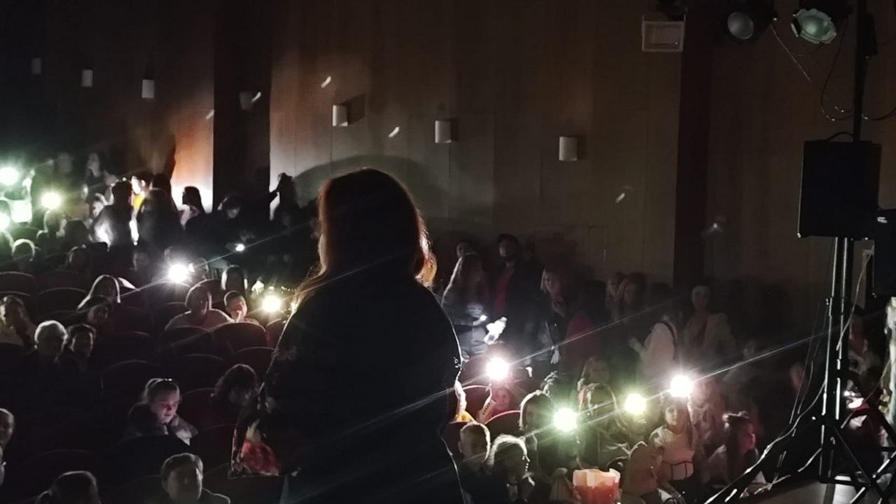 Темна фотографія з концертної зали або кінотеатру, де жінка стоїть на сцені спиною до глядачів. Аудиторія (переважно молодь) сидить у червоних кріслах і освітлює сцену та залу ліхтариками мобільних телефонів, створюючи багато яскравих точок світла.