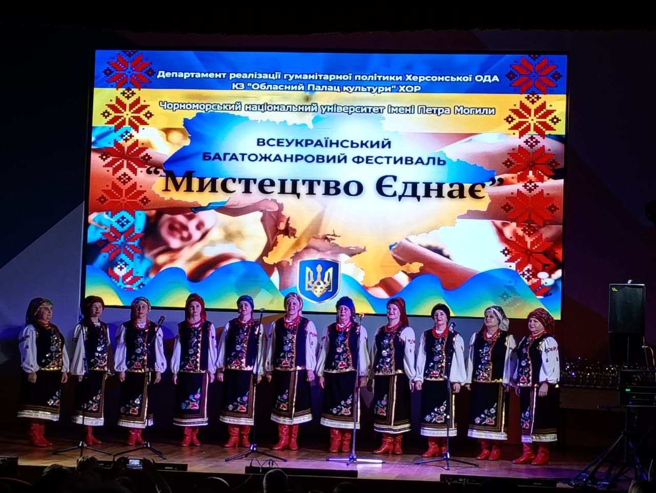 На сцені виступає жіночий хоровий колектив (11 учасниць) у традиційному українському вбранні (вишиванки, жилетки та червоні чоботи). Над ними розташований великий екран із синьо-жовтим фоном, на якому великими літерами написано: ВСЕУКРАЇНСЬКИЙ БАГАТОЖАНРОВИЙ ФЕСТИВАЛЬ МИСТЕЦТВО ЄДНАЄ.