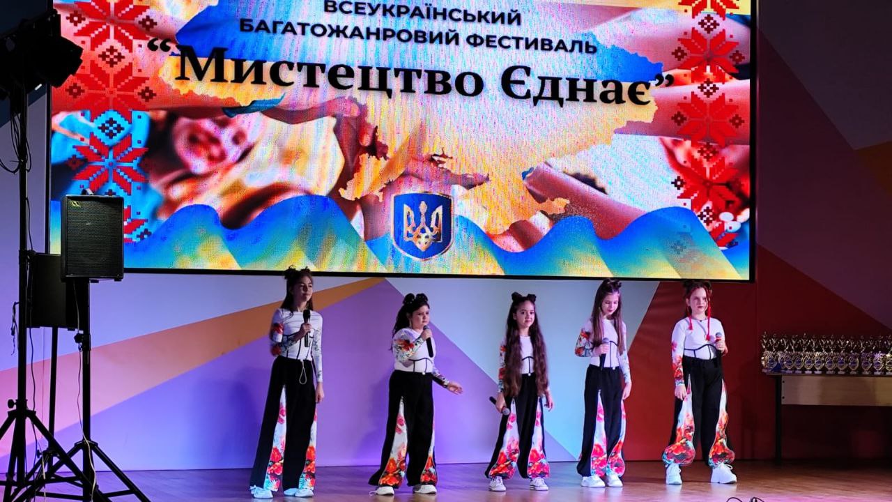 П'ятеро молодих дівчат виступають на сцені у сучасних чорно-білих костюмах з квітковими візерунками, тримаючи мікрофони. Позаду них розташований великий екран із синьо-жовтим фоном та написом ВСЕУКРАЇНСЬКИЙ БАГАТОЖАНРОВИЙ ФЕСТИВАЛЬ 