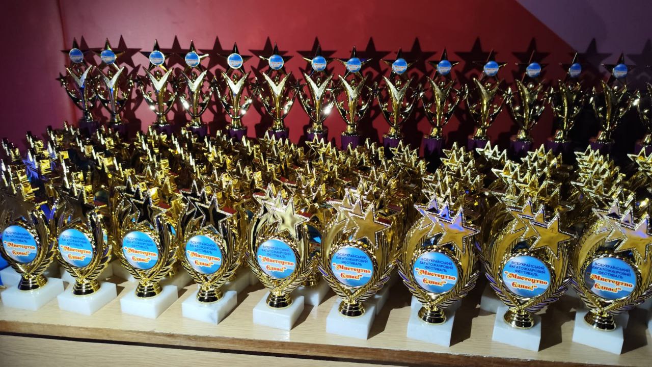 Два ряди численних золотих нагородних кубків та призів у формі зірок, виставлених на дерев'яній полиці на тлі червоної стіни. На медальйонах кубків написано: ВСЕУКРАЇНСЬКИЙ БАГАТОЖАНРОВИЙ ФЕСТИВАЛЬ 