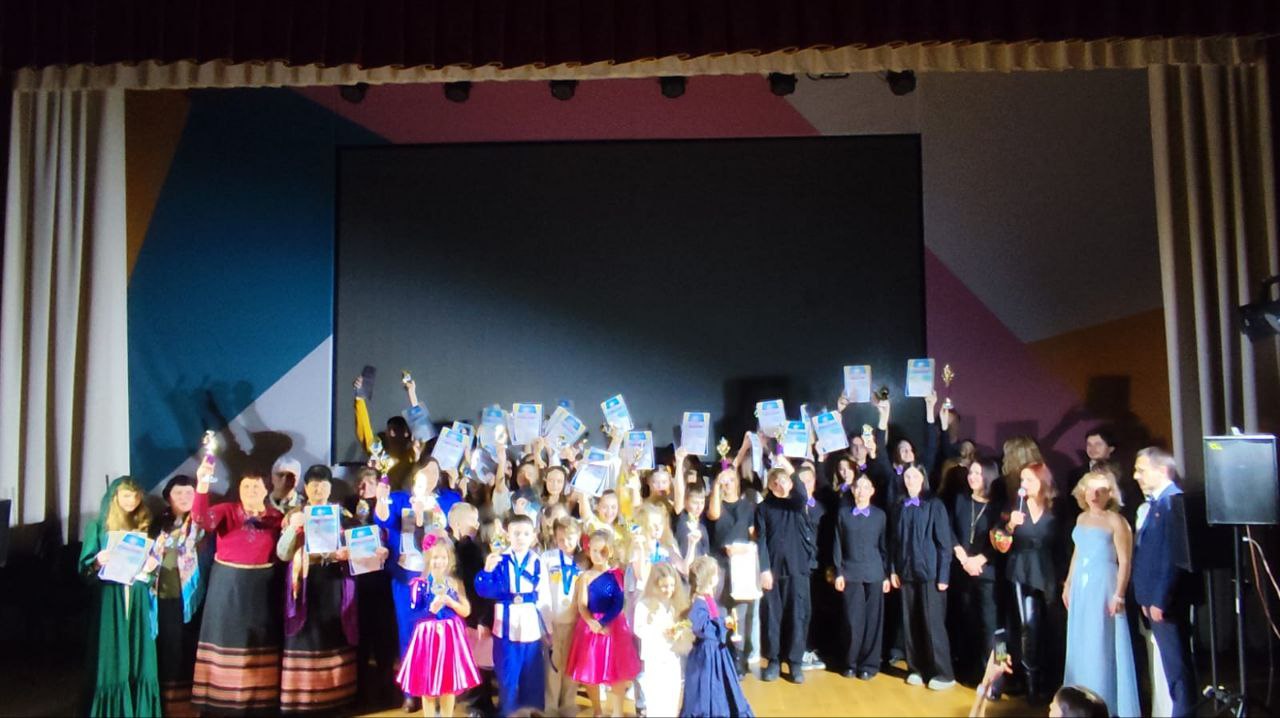 Велика група учасників та організаторів, включаючи дітей у сценічних костюмах і дорослих, стоїть на сцені після виступу, тримаючи високо над головою нагороди, дипломи та сертифікати. Позаду них — великий темний екран та завіса.