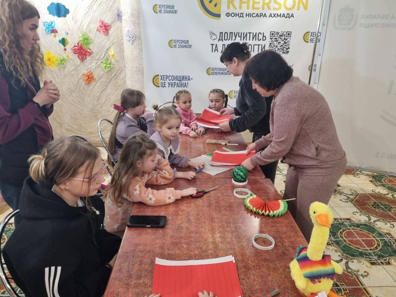 Група дівчаток різного віку сидить за столом і займається рукоділлям, імовірно, створюючи прикраси з червоного паперу, схожі на кавун, під керівництвом двох дорослих жінок. На стінах видно плакати з написами українською мовою, зокрема: ХЕРСОНЩИНА - ЦЕ УКРАЇНА! та ХЕРСОНЦІВ НЕ ЗЛАМАТИ! На столі лежать ножиці, скотч, папір і іграшка-каченя.