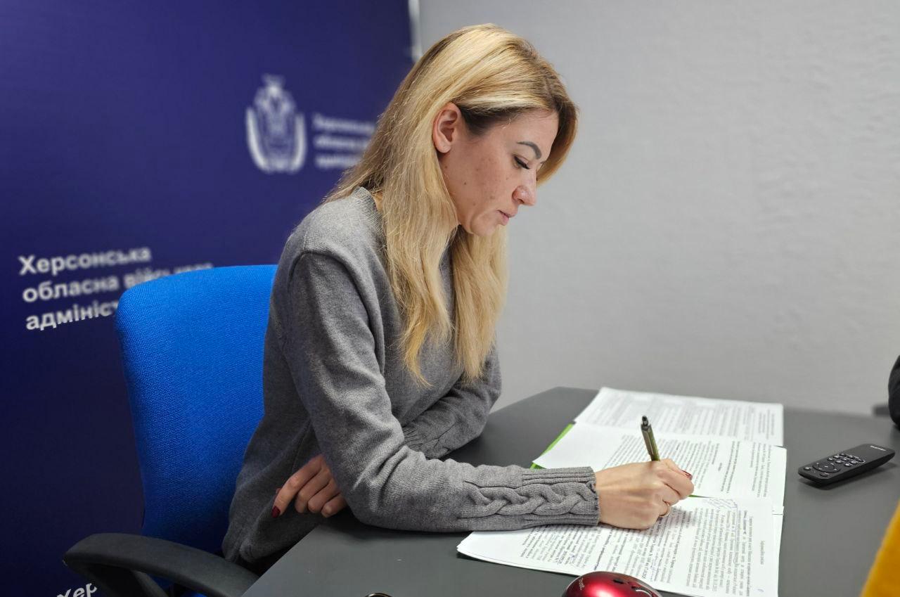 Жінка у сірому светрі сидить за робочим столом на фоні синього банера з логотипом Херсонської обласної військової адміністрації. Вона зосереджено робить записи ручкою у документах, що лежать перед нею на столі.