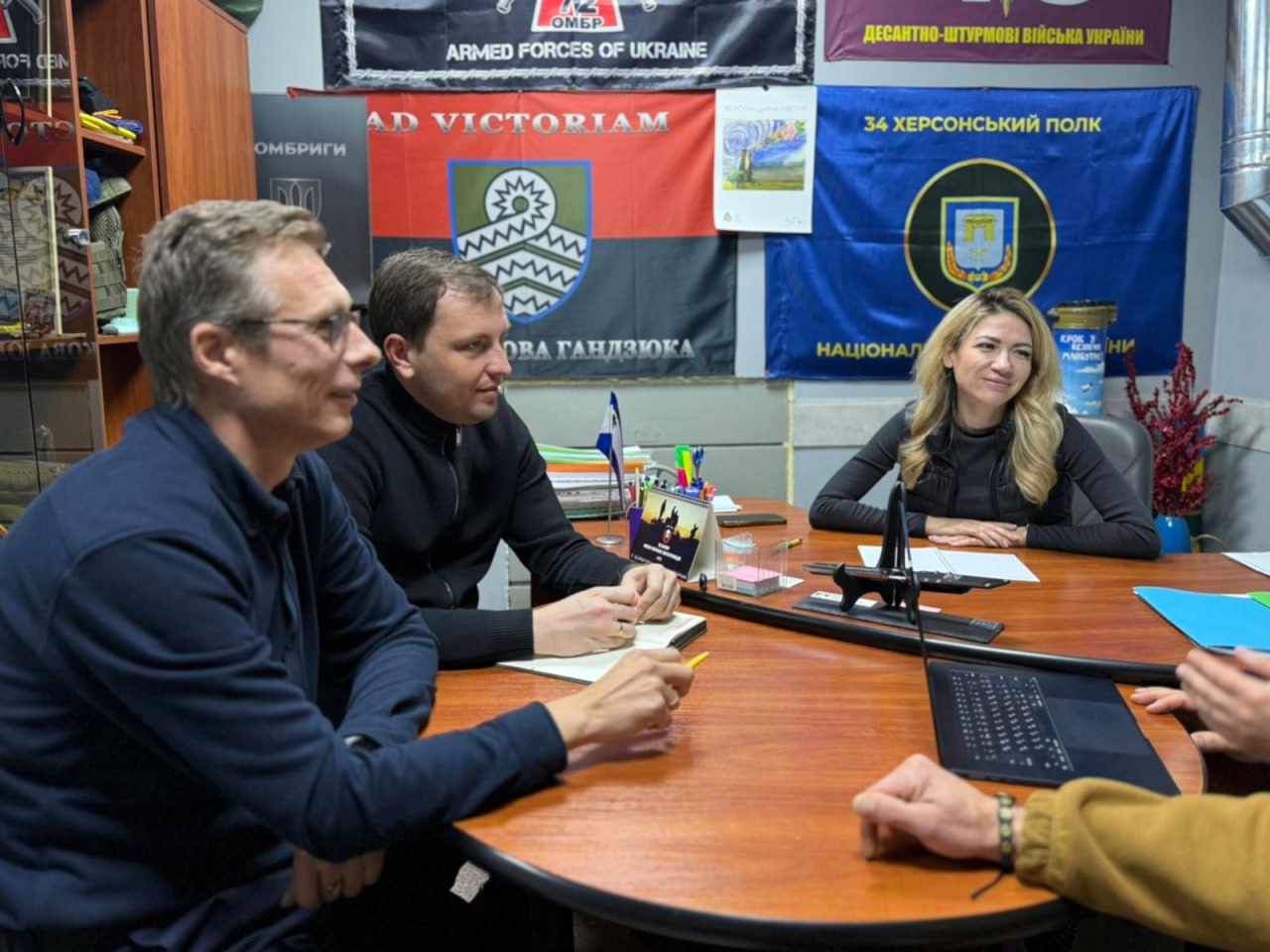 Троє людей (двоє чоловіків і одна жінка) сидять за круглим столом під час зустрічі. На стіні позаду них висять військові прапори, зокрема прапор 34-го Херсонського полку та прапор 59 ОШБр ім. Якова Гандзюка із написом: AD VICTORIAM.