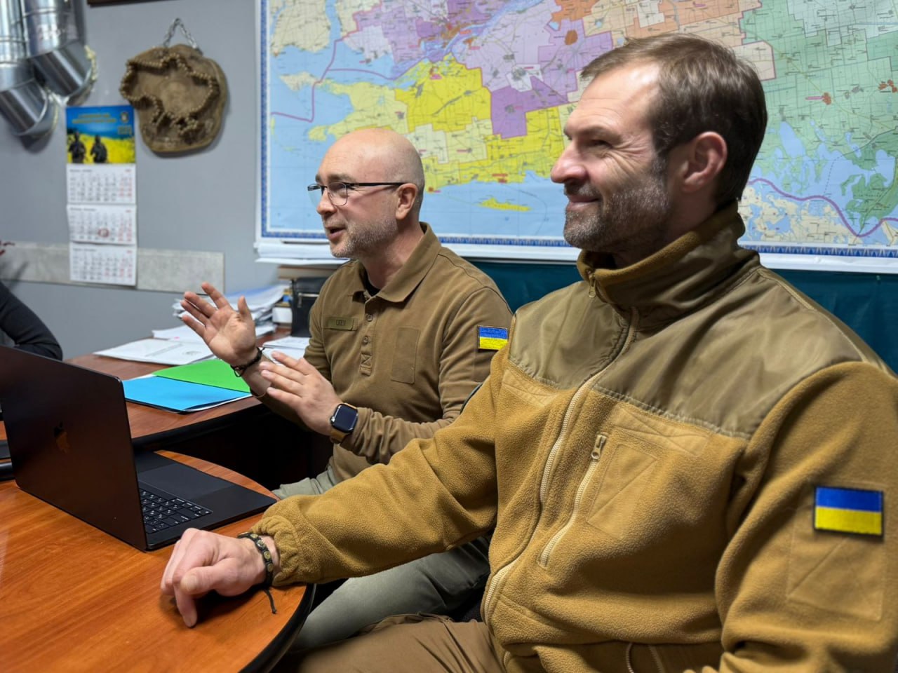 Двоє чоловіків у військовій формі (один у коричневій флісовій куртці з прапором України на рукаві, інший у сорочці поло) сидять за столом перед ноутбуком. Чоловік зліва активно жестикулює під час розмови. На стіні позаду них висить велика карта.