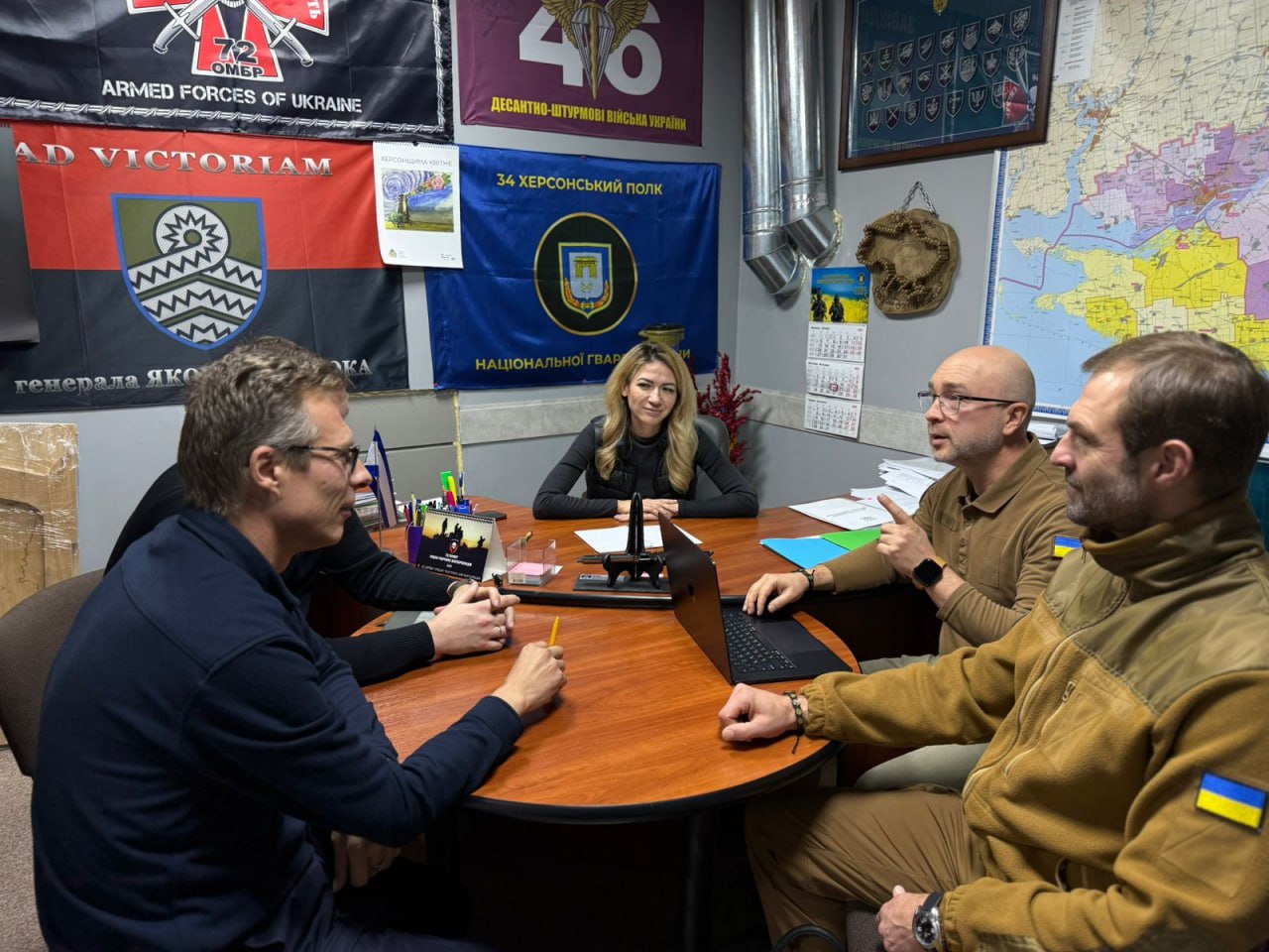 Четверо людей (троє чоловіків і одна жінка) сидять за круглим столом, обговорюючи щось на зустрічі. На стіні позаду них висять військові прапори різних підрозділів, включаючи 34-й Херсонський полк, а також велика мапа.