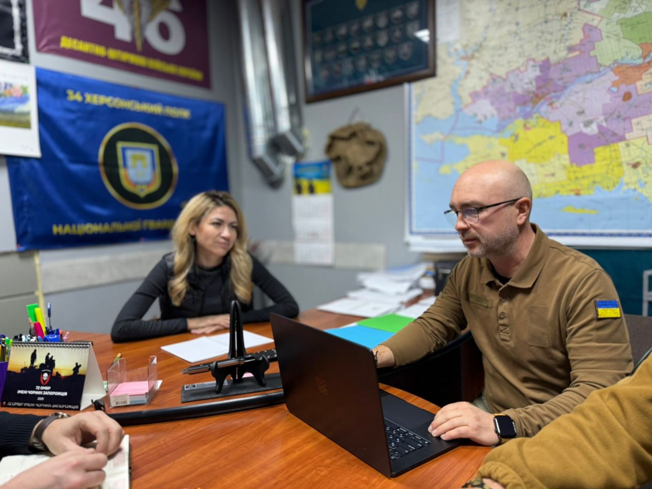Чоловік у військовій формі з прапором України на рукаві працює за ноутбуком під час зустрічі. Жінка зі світлим волоссям сидить навпроти, слухаючи його. На задньому плані видно прапор 34-го Херсонського полку та мапу.