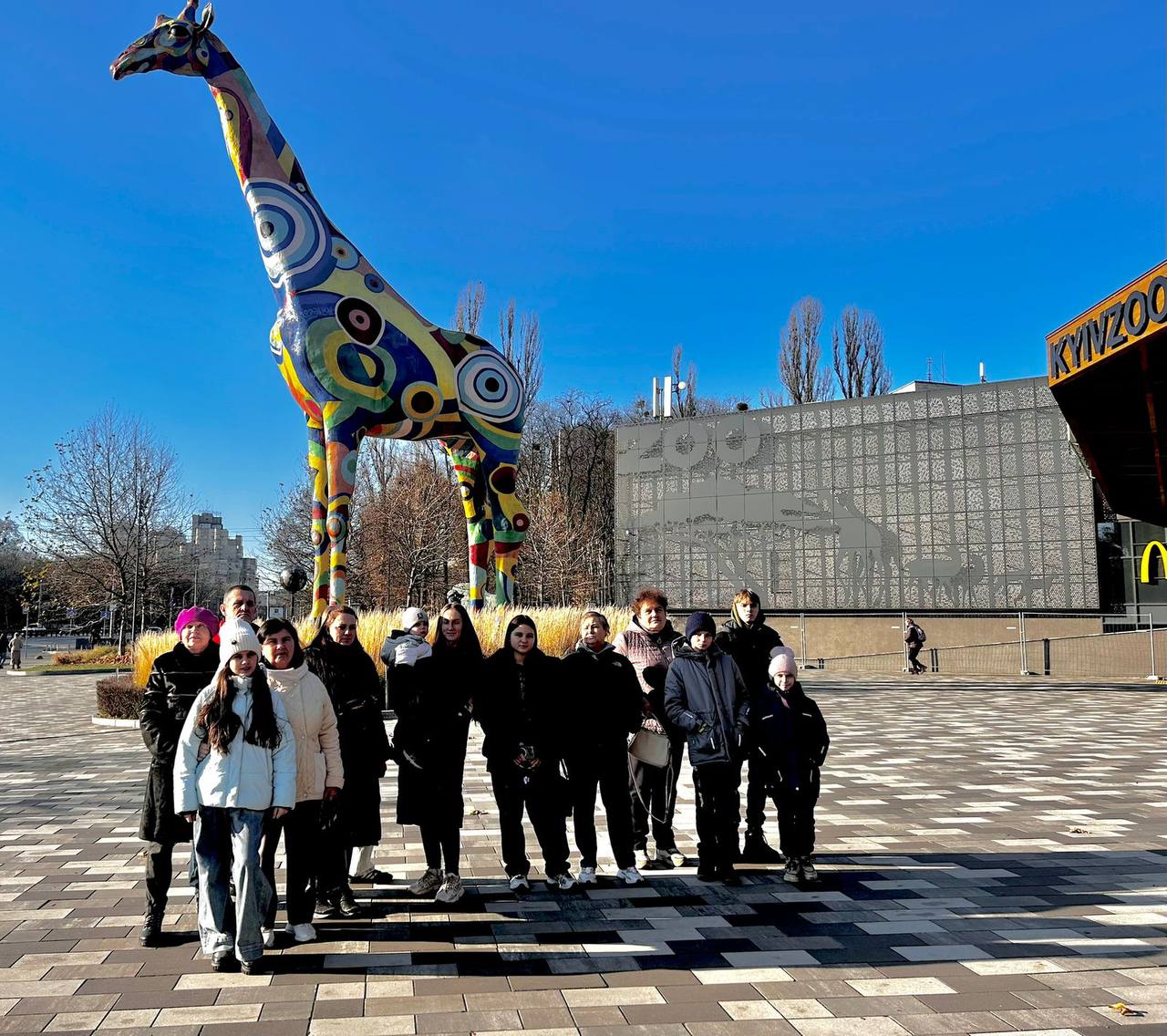 Група підлітків і молоді стоїть на площі перед входом до Київського зоопарку (КИЇВЗОО) у сонячний день. На задньому плані домінує велика, яскраво розфарбована скульптура жирафа, прикрашена різнокольоровими геометричними та органічними візерунками. Скульптура розташована перед сучасною будівлею зоопарку.