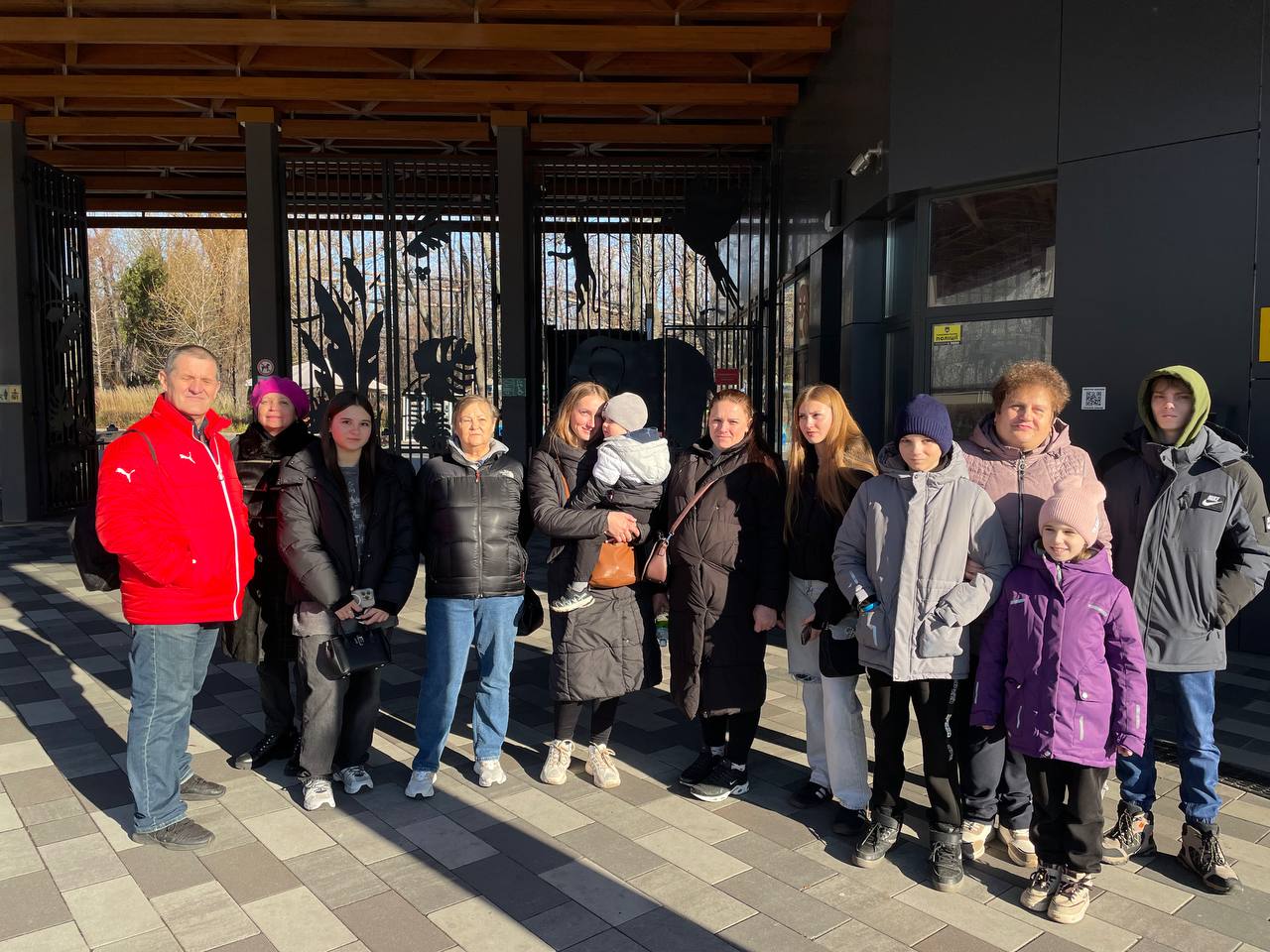 Група з приблизно десяти дорослих, підлітків і дітей, одягнених у зимовий одяг, стоїть і позує для фото на сонячній площі перед великими металевими воротами, ймовірно, входу до зоопарку. Чоловік ліворуч одягнений у яскраву червону куртку, а жінка в центрі тримає на руках маленьку дитину.