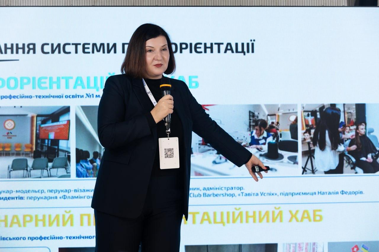Жінка в чорному діловому костюмі виступає на презентації, тримаючи мікрофон і вказуючи рукою на екран за спиною. На екрані видно текст про профорієнтацію та дві невеликі фотографії, пов'язані з перукарською справою.