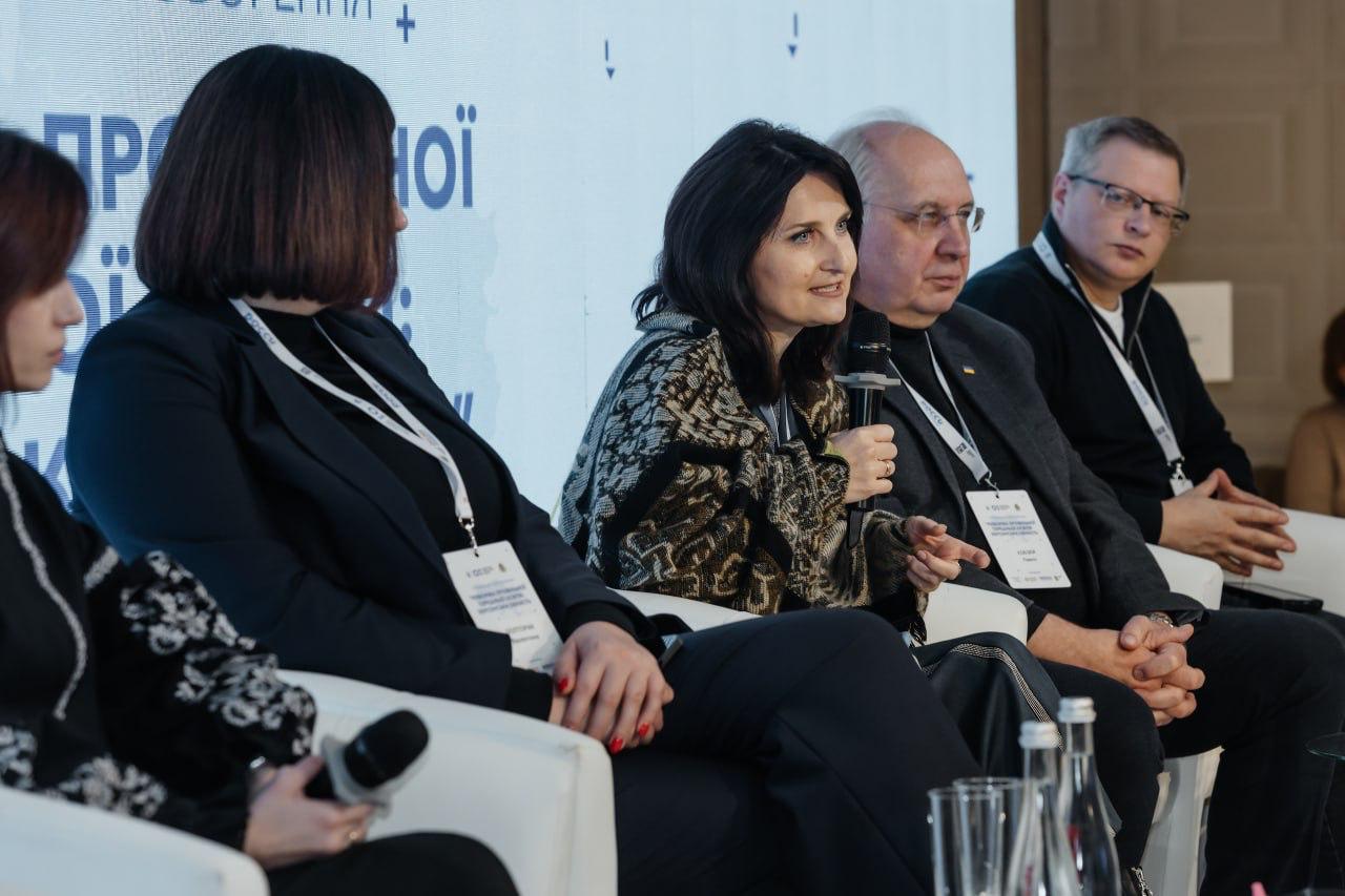 Група людей сидить на сцені в білих кріслах під час панельної дискусії. Жінка в центрі, одягнена у візерунчастий светр, говорить у мікрофон, поруч із нею сидять двоє чоловіків та ще одна жінка.