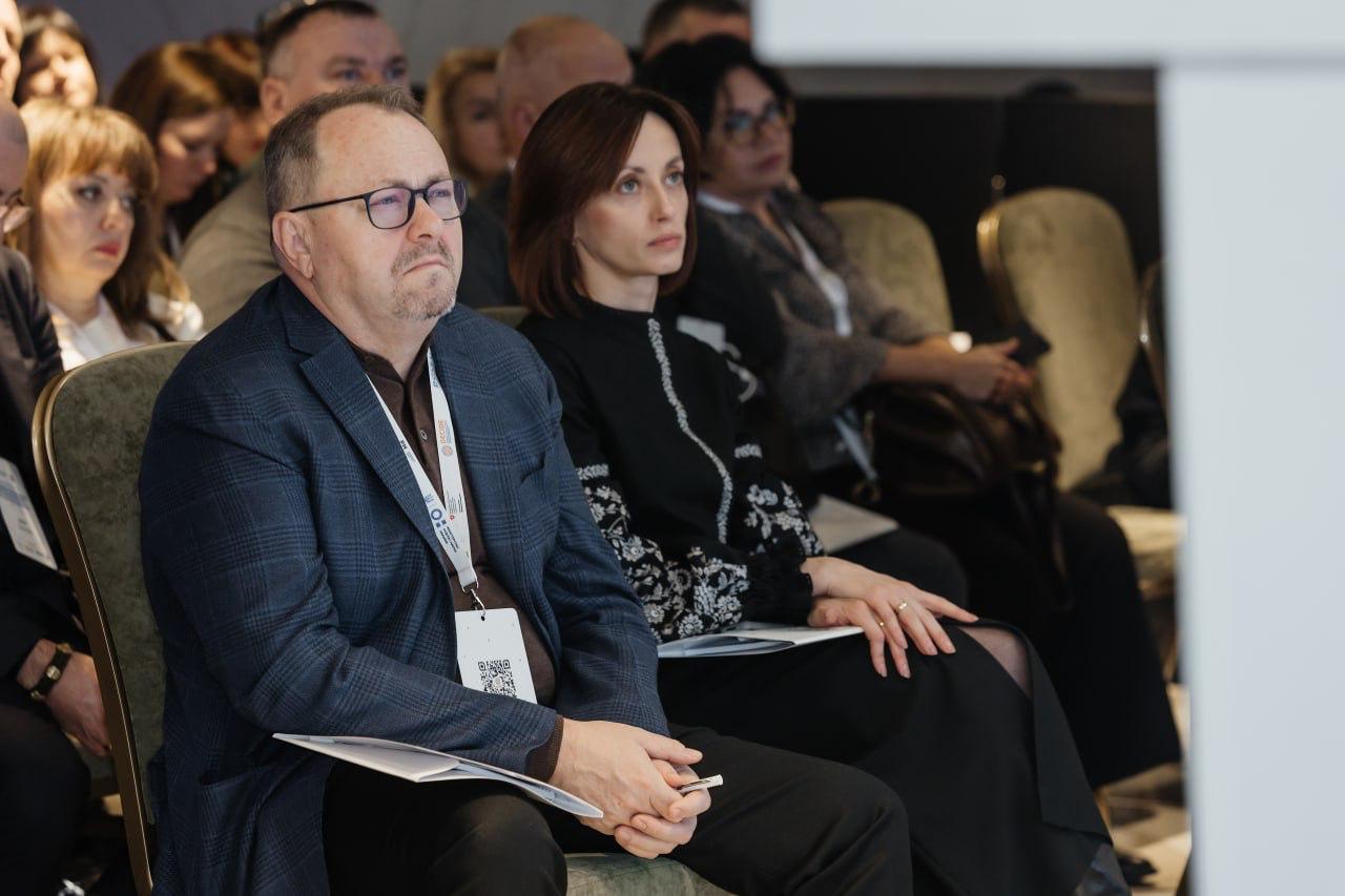 Чоловік у синьому піджаку та окулярах і жінка у чорній вишиванці уважно слухають, сидячи в першому ряду на конференції. Обидва тримають папери, а на задньому плані видно інших учасників заходу.