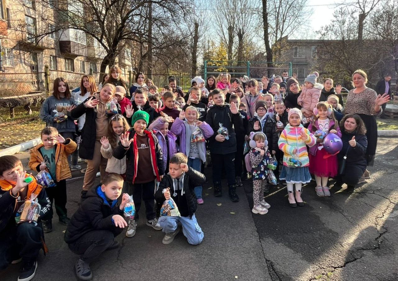  Велика група усміхнених дітей різного віку та кількох дорослих стоять надворі, ймовірно, у дворі житлового будинку, та махають руками, тримаючи в руках пакети з подарунками або солодощами.