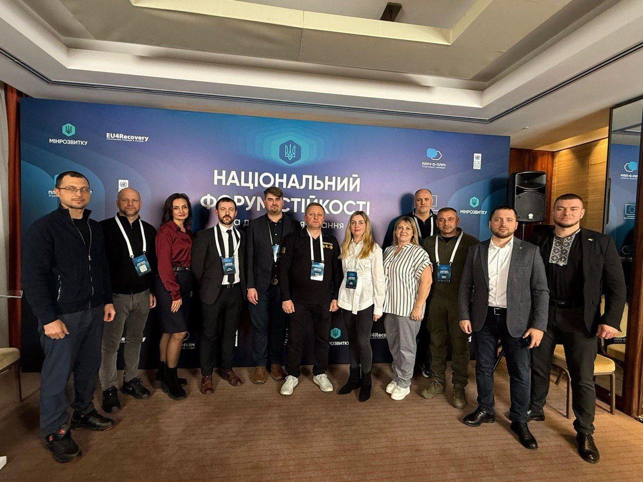 Велика група з одинадцяти осіб стоїть перед банером НАЦІОНАЛЬНИЙ ФОРУМ СТІЙКОСТІ. На банері також видно логотипи МІНРЕЗВИТКУ, EU4Recovery, Пліч-о-пліч та інші. Усі учасники одягнені у діловий або повсякденний одяг та мають бейджики.