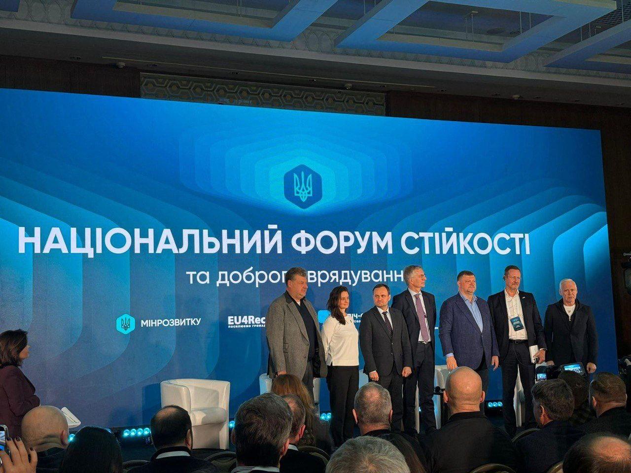 Група з семи чоловіків та однієї жінки стоїть на сцені під час НАЦІОНАЛЬНОГО ФОРУМУ СТІЙКОСТІ та доброго врядування. За ними — великий синій екран з логотипами, зокрема МІНРЕЗВИТКУ та EU4Rec. На передньому плані видно частину аудиторії.