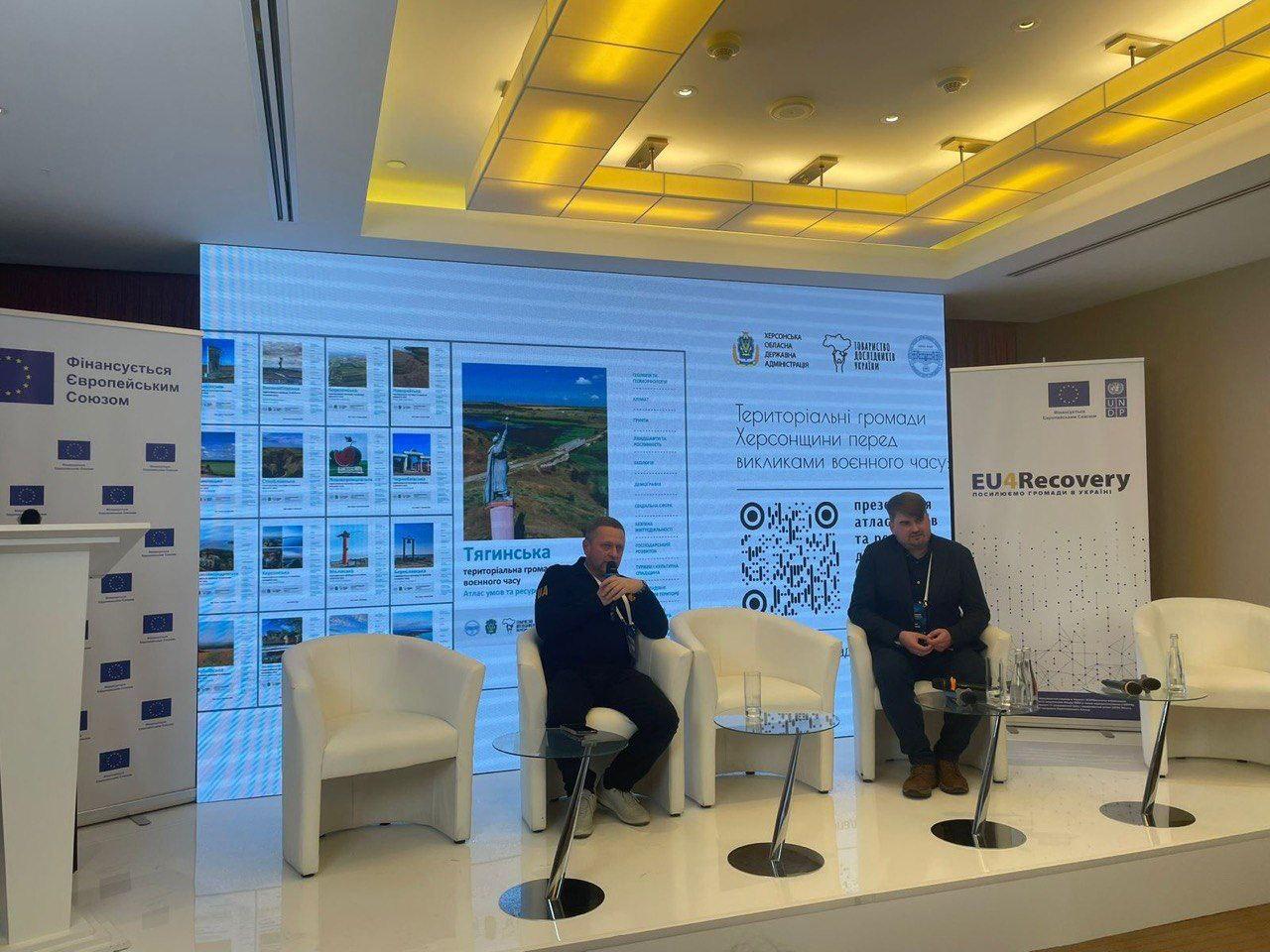 Двоє чоловіків сидять у кріслах на сцені, обговорюючи Територіальні громади Херсонщини перед викликами воєнного часу. На екрані видно назву Тягинська та логотип EU4Recovery.