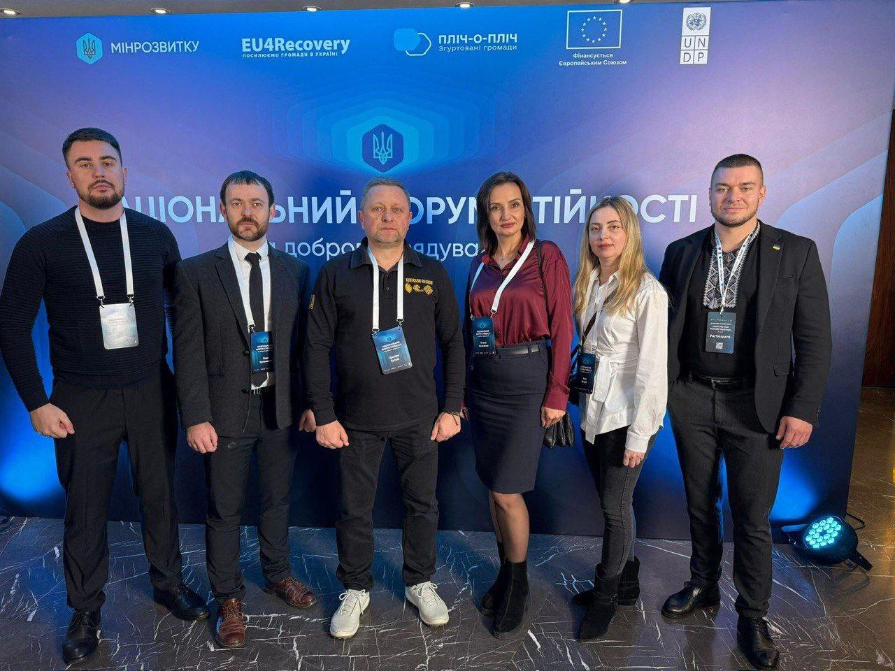 Шестеро людей (п'ятеро чоловіків та одна жінка) стоять перед синім банером із написами: МІНРЕЗВИТКУ, EU4Recovery, Пліч-о-пліч, логотипами ЄС та ООН (UNDP). На банері видно великий напис, частина якого: НАЦІОНАЛЬНИЙ ФОРУМ СТІЙКОСТІ. Усі учасники мають на шиї ідентифікаційні картки.