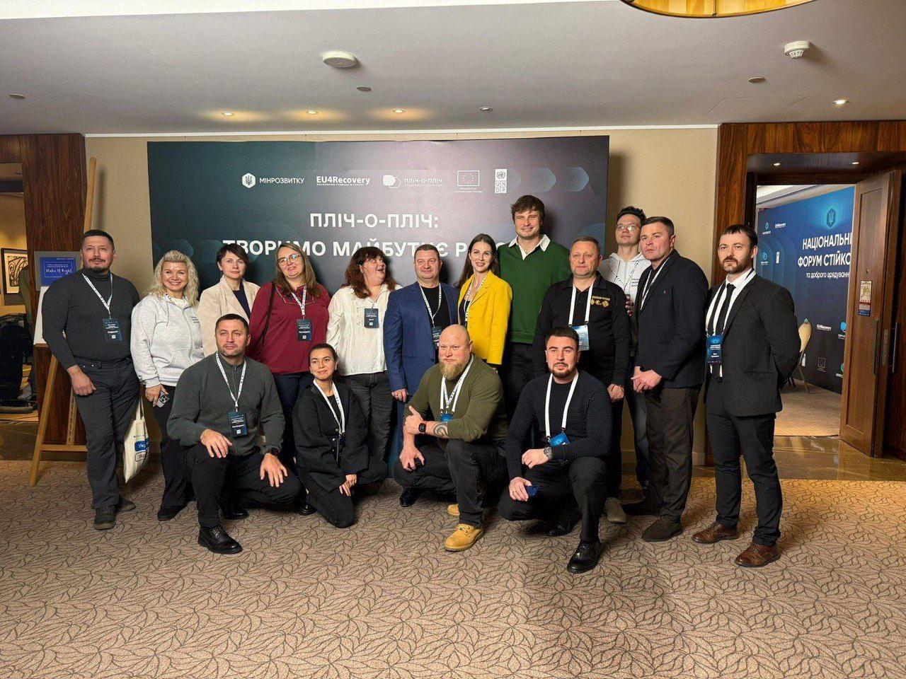 Групове фото п'ятнадцяти осіб (чотирнадцяти дорослих та однієї дитини) перед банером події з логотипами Мінрегіону (Міністерства розвитку громад та територій України) та EU4Recovery. На банері написано: ПЛІЧ-О-ПЛІЧ: СТВОРЮЄМО МАЙБУТНЄ РАЗОМ.