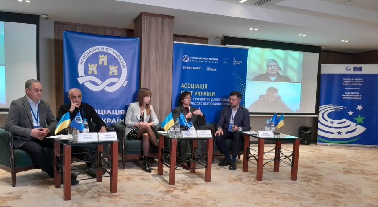 П'ятеро учасників сидять на панельній дискусії на заході Асоціації міст України. Вони розташовані на сцені перед синіми банерами з логотипом організації. На столі перед ними стоять українські прапорці. На екрані позаду них видно двох додаткових учасників, які приєдналися онлайн через відеозв'язок.