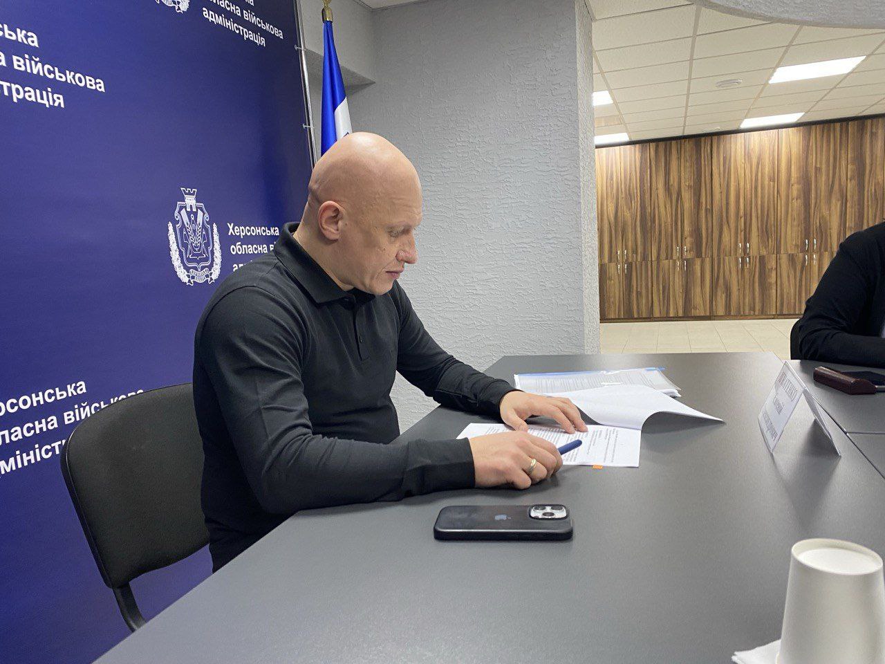 Чоловік середнього віку з лисою головою, одягнений у чорну сорочку поло, сидить за столом під час наради. Він уважно читає або підписує документи, тримаючи в руці ручку. На задньому плані видно синій банер із текстом: Херсонська обласна військова адміністрація.