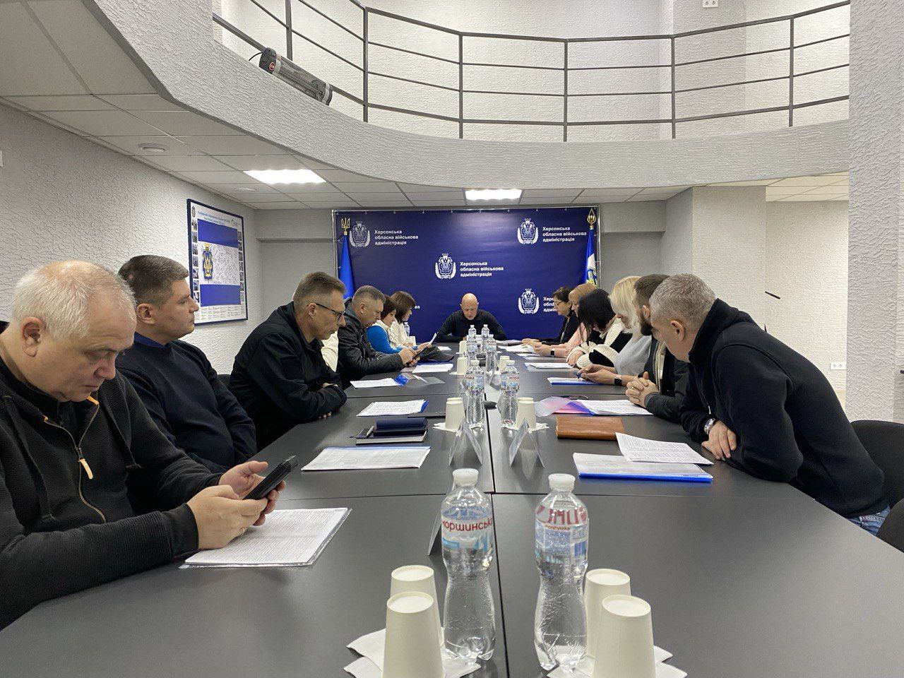 Група з приблизно десяти-дванадцяти людей, більшість із яких чоловіки та жінки в діловому або повсякденному одязі, сидять за довгим столом у кімнаті для нарад. На задньому плані розташований темно-синій банер із символікою та назвою: Херсонська обласна державна адміністрація. На столі лежать документи, телефони та пляшки з водою.