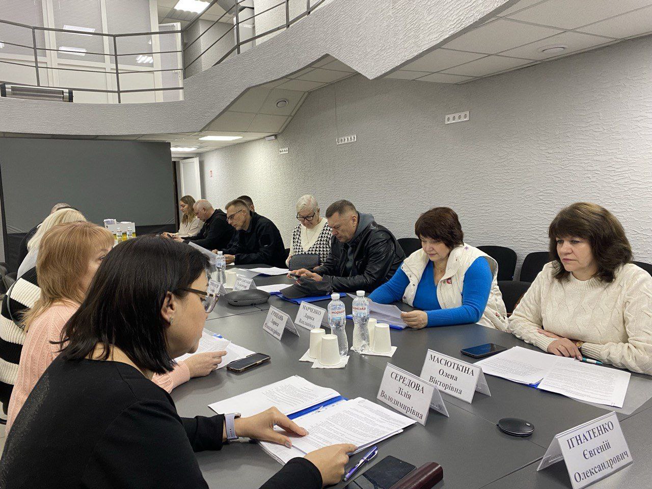 Група людей, включно з чоловіками та жінками, сидять навколо великого столу під час наради. Перед ними розкладені документи, таблички з іменами та пляшки з водою. Жінка на передньому плані в окулярах переглядає папери.